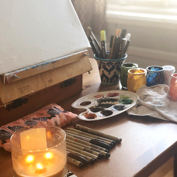 Artist supplies. There's a dim candle, paint palette, a canvas, and some brushes.